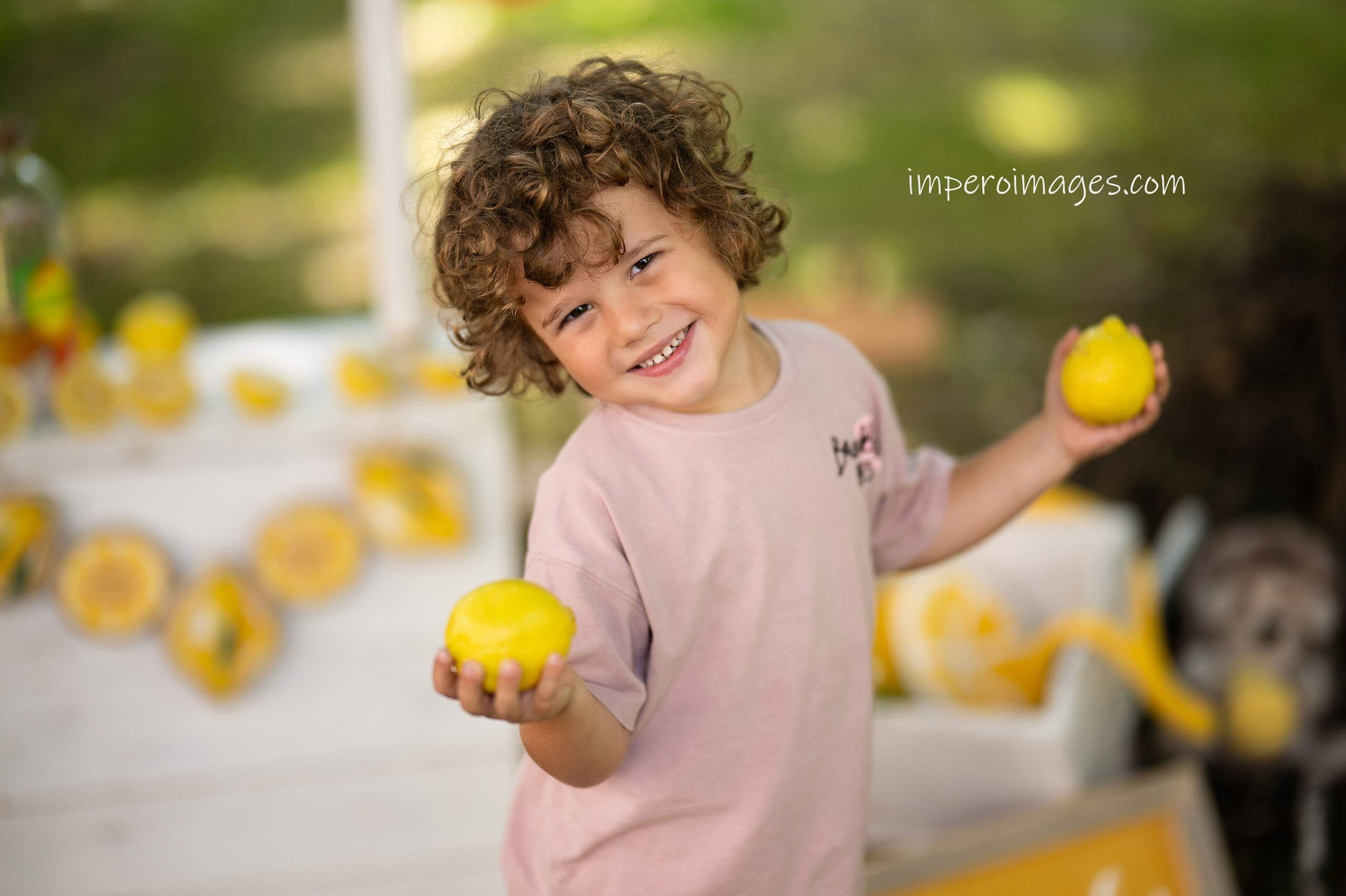 Lemoniadowa sesja przedszkolna na zakończenie roku — uśmiechnięty chłopiec z kręconymi włosami trzymający cytryny w słonecznym plenerze
