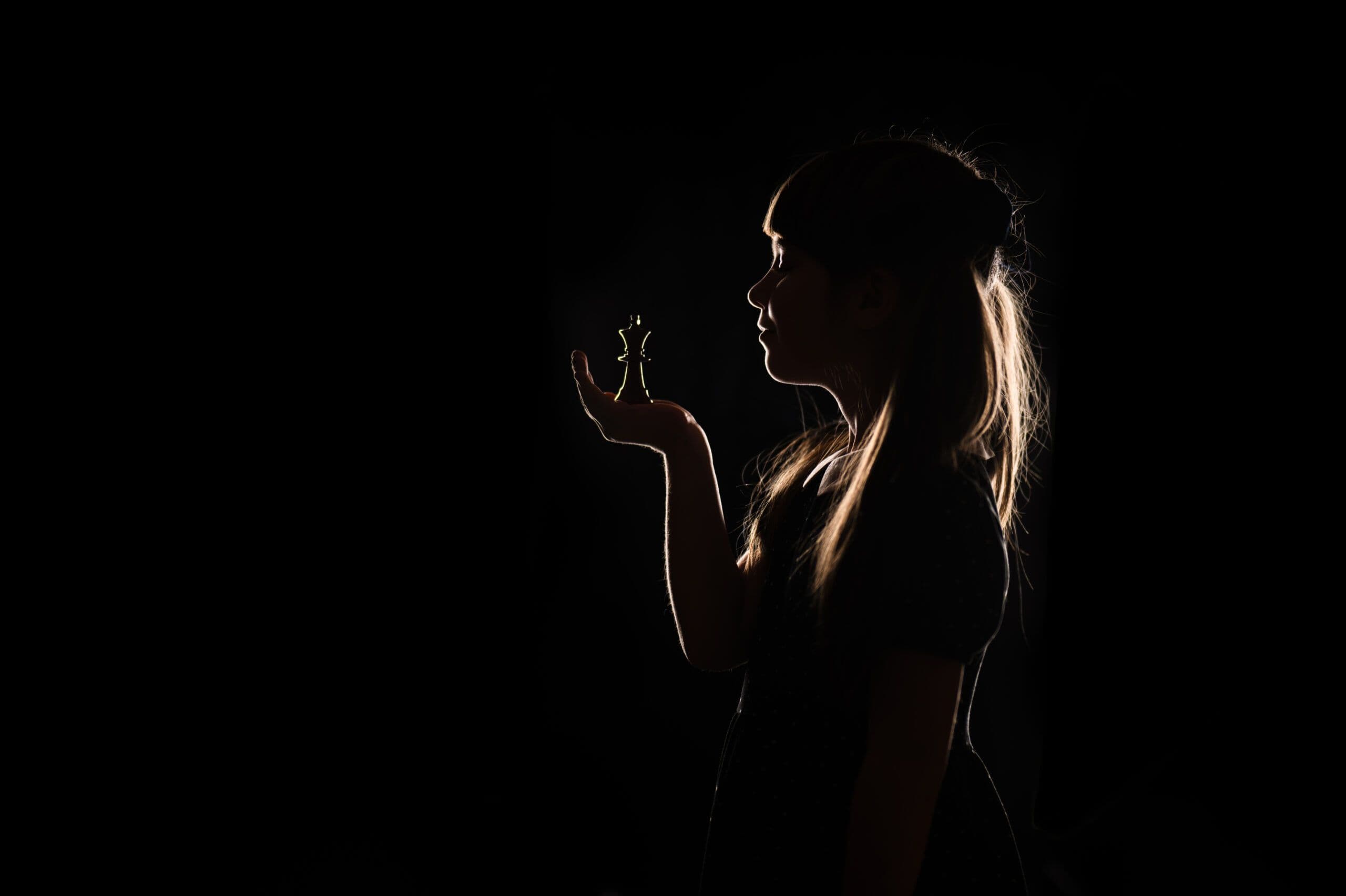 Artystyczny portret szkolny malowany światłem — dziewczynka z figurą hetmana szachowego w sylwetce na ciemnym tle „Królowa z Królową"