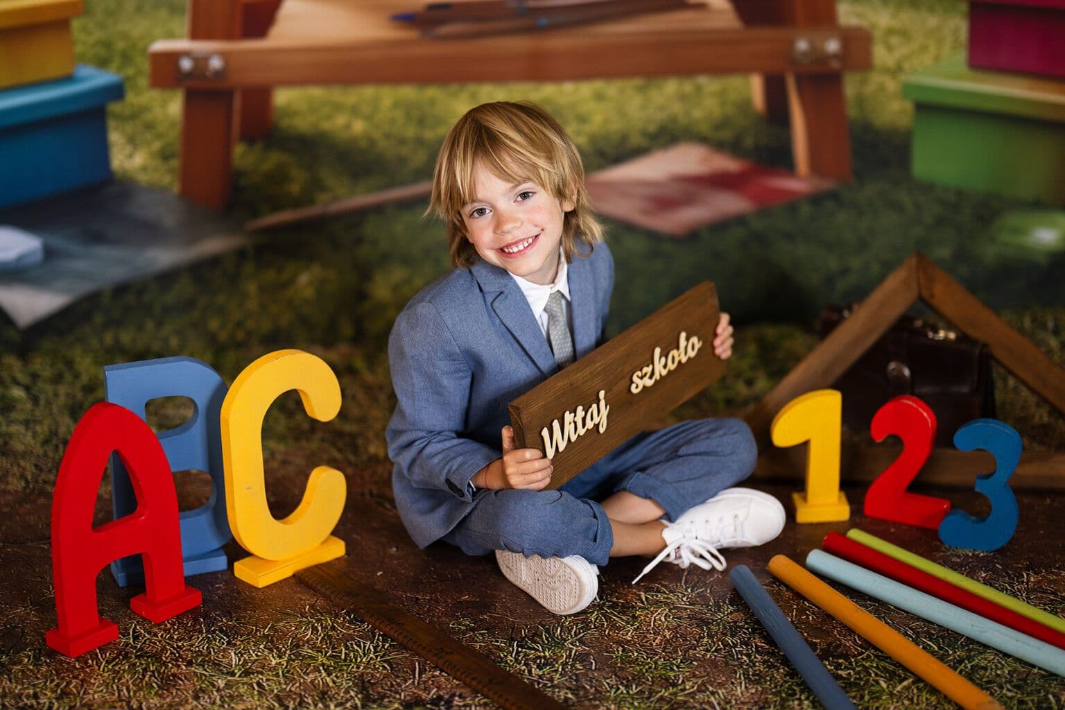 Sesja pasowania na ucznia — uśmiechnięty chłopiec w eleganckim garniturze z tabliczką „Witaj szkoło" wśród kolorowych liter ABC i cyfr 123