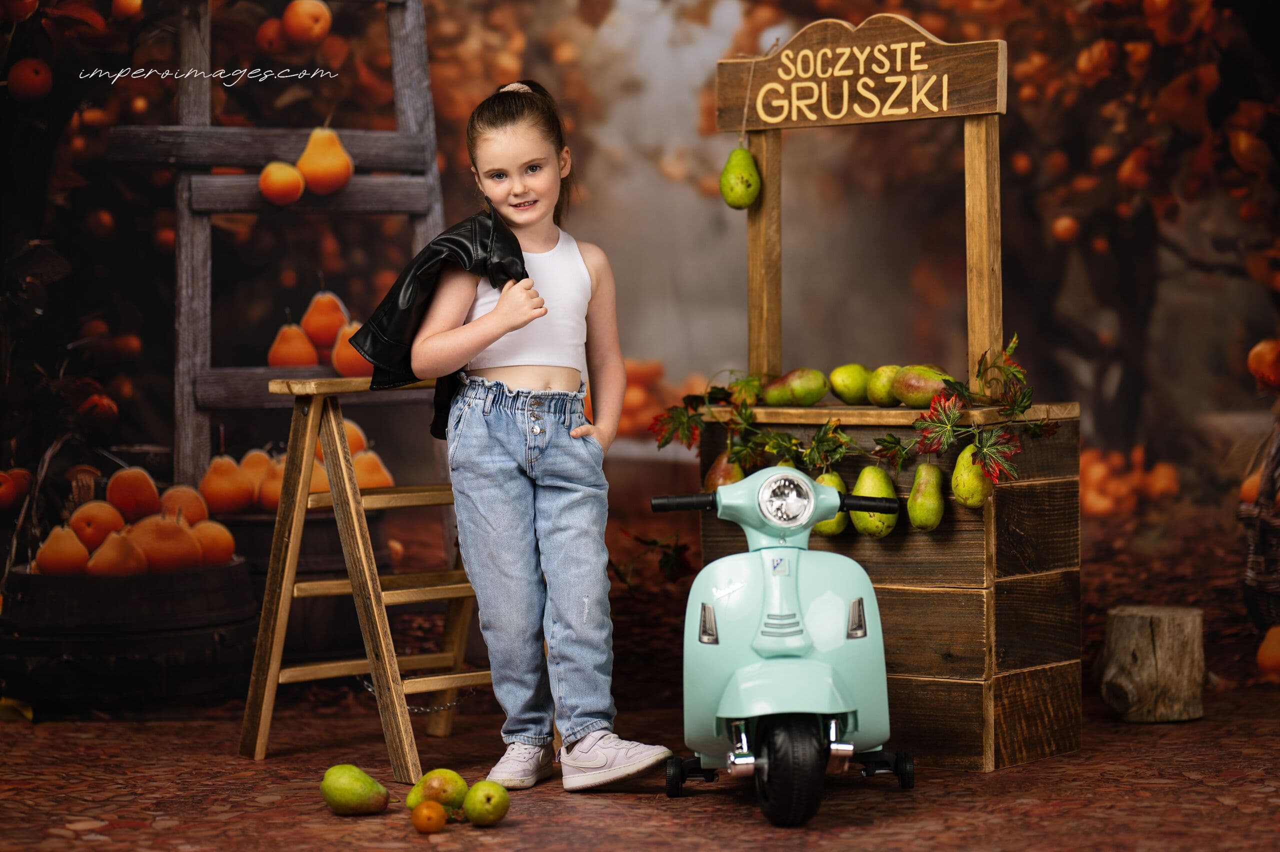 Jesienna sesja przedszkolna — dziewczynka przy skuterze Vespa i drewnianym straganiku z gruszkami w rustykalnej scenerii