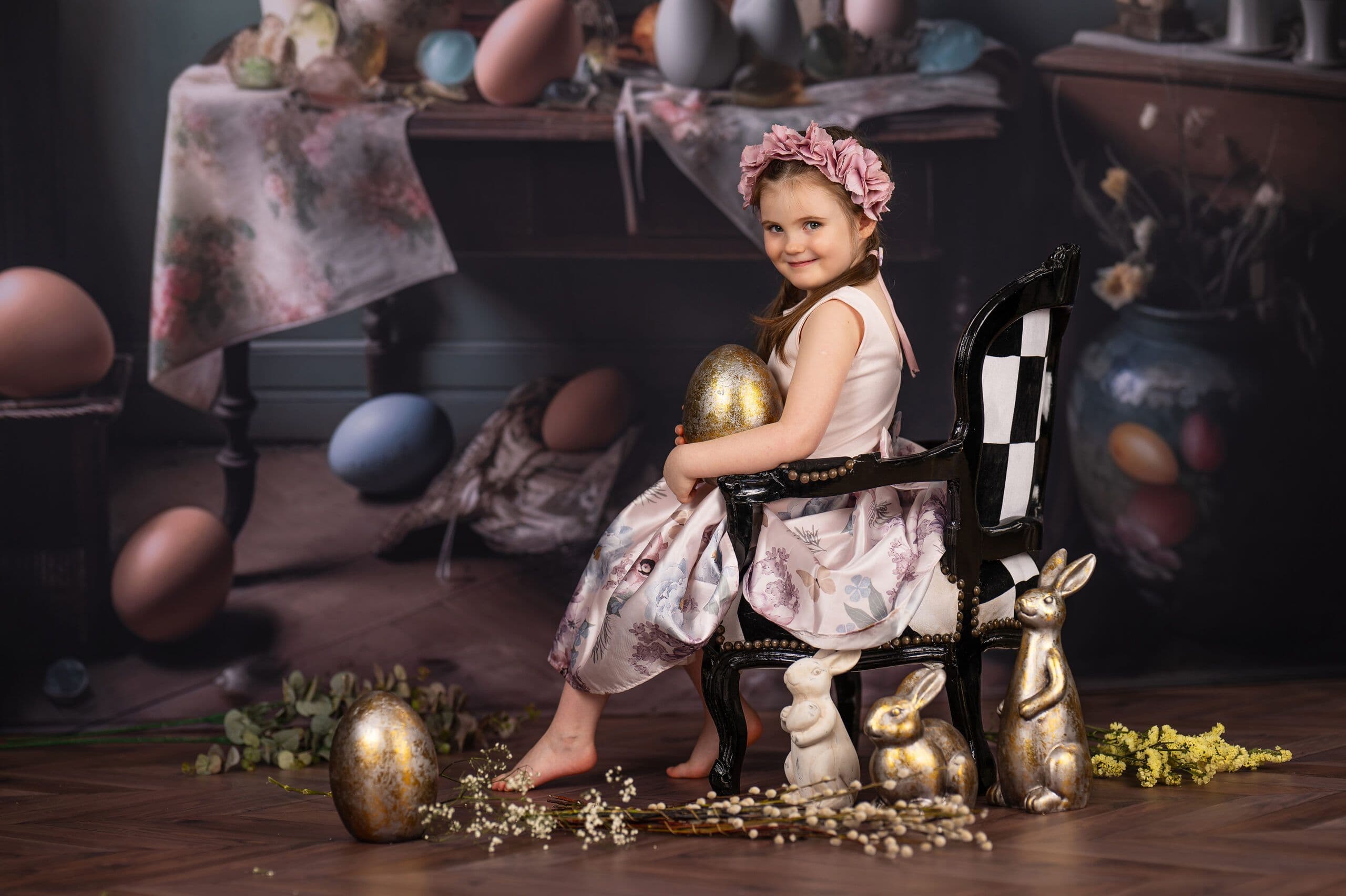 Wielkanocna sesja w stylu Alicji w Krainie Czarów — dziewczynka z wiankiem kwiatowym na szachowym fotelu ze złotymi pisankami i dekoracyjnymi zajączkami